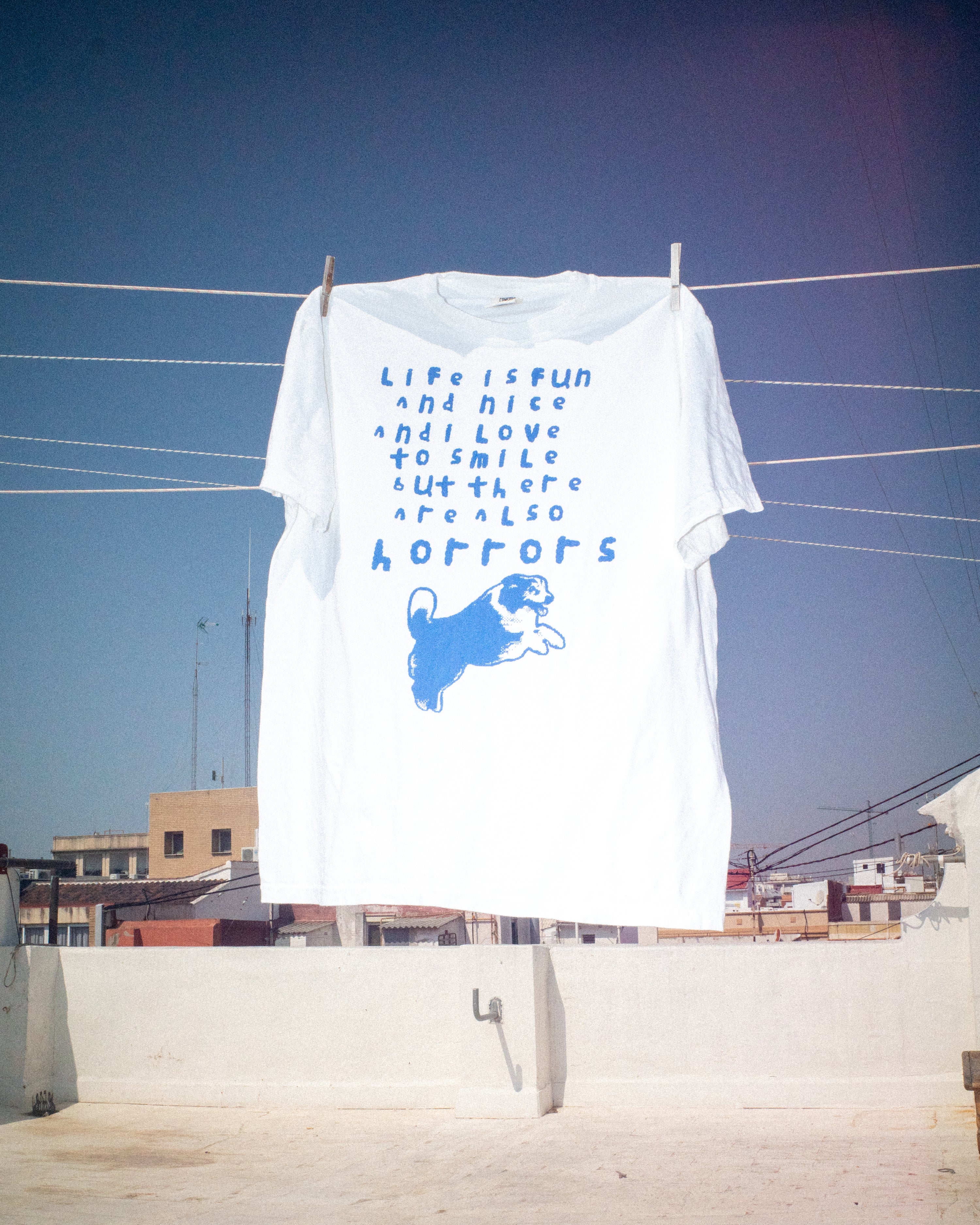 White t-shirt with blue text and dog illustration on a clothesline against a clear blue sky.
